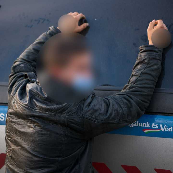 Letartóztatás, kitakart arcú férfi rendőrautóhoz támaszkodva vár